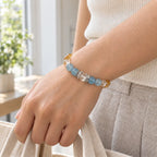 Woman wearing a citrine and aquamarine bracelet while holding a cream canvas tote beside a bright window.